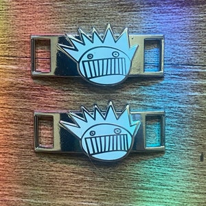 May include: Two silver shoe charms featuring a cartoon face design. The face is white with black details, including a spiky crown, eyes, and a mouth with vertical lines. The charms are rectangular with a slot for attaching to shoes.