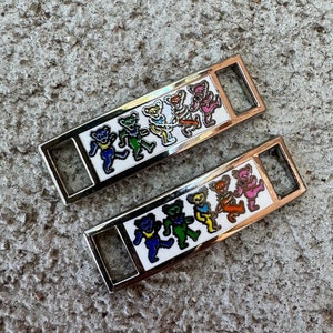 May include: Two silver metal shoe charms with a white background and a design of six dancing bears in different colors. The bears are wearing different colored hats and have different colored bodies.