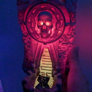 Puede incluir: Una escultura detallada e iluminada que presenta una calavera dentro de un marco circular, con una escalera que conduce a un dado. La escultura est&aacute; iluminada con tonos rojos y naranjas, creando un efecto espeluznante. El dise&ntilde;o evoca un tema de fantas&iacute;a o g&oacute;tico.