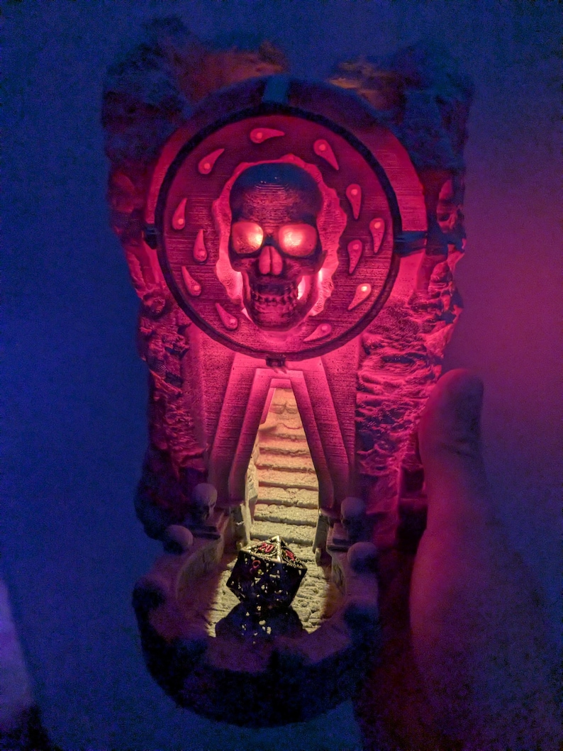 Puede incluir: Una escultura detallada e iluminada que presenta una calavera dentro de un marco circular, con una escalera que conduce a un dado. La escultura est&aacute; iluminada con tonos rojos y naranjas, creando un efecto espeluznante. El dise&ntilde;o evoca un tema de fantas&iacute;a o g&oacute;tico.