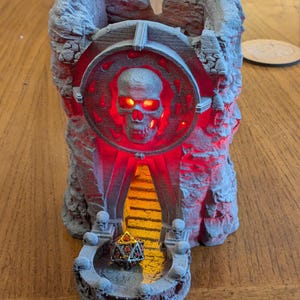 Puede incluir: Una torre de dados gris, impresa en 3D, con forma de ruina de castillo, con un dise&ntilde;o de calavera y ojos rojos brillantes e iluminaci&oacute;n interior. Un dado de m&uacute;ltiples caras descansa en la base. La torre est&aacute; dise&ntilde;ada para juegos de mesa.