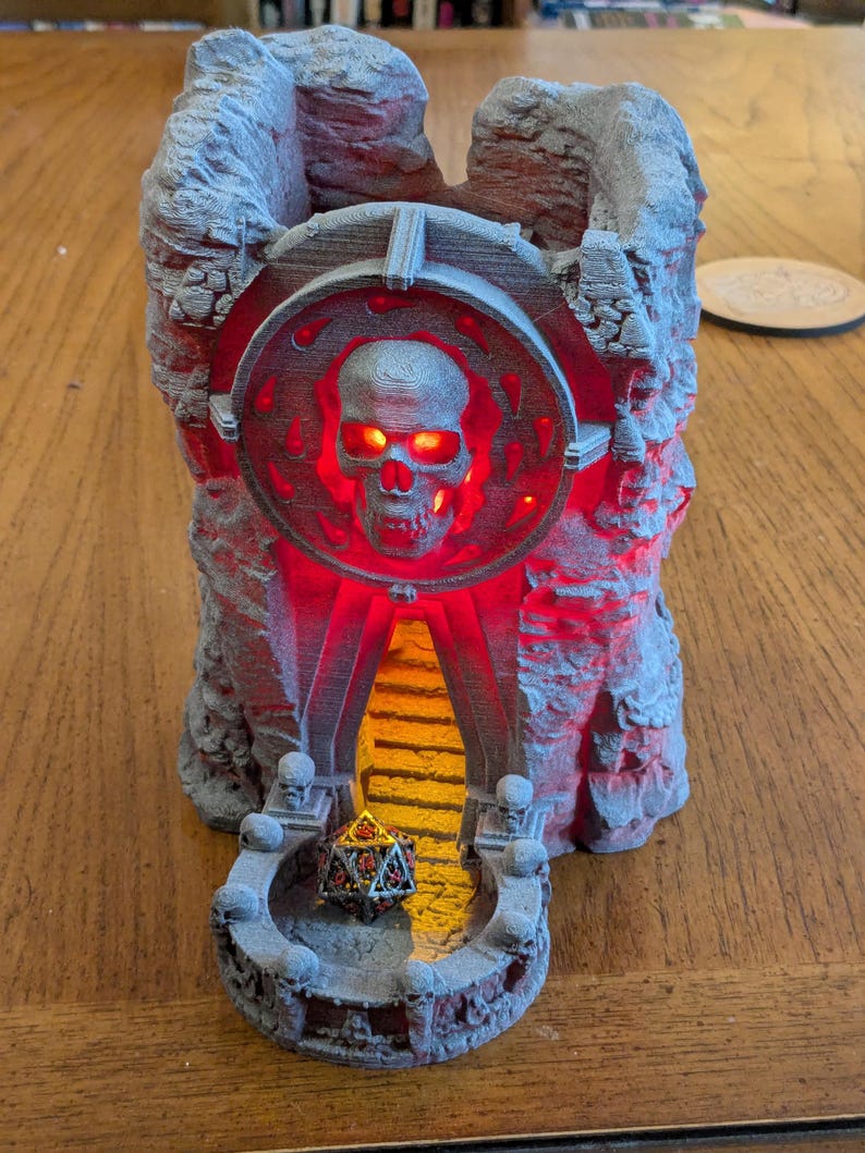 Puede incluir: Una torre de dados gris, impresa en 3D, con forma de ruina de castillo, con un dise&ntilde;o de calavera y ojos rojos brillantes e iluminaci&oacute;n interior. Un dado de m&uacute;ltiples caras descansa en la base. La torre est&aacute; dise&ntilde;ada para juegos de mesa.