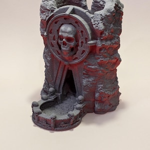 Puede incluir: Una torre de dados gris, impresa en 3D, con un dise&ntilde;o de calavera. La torre presenta una calavera en el centro, rodeada por un marco circular. La base tiene peque&ntilde;os detalles de calavera. La estructura tiene una apariencia rocosa, similar a una cueva.
