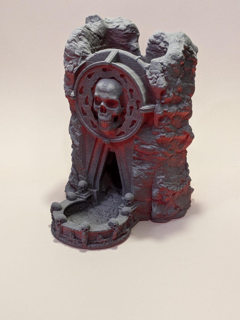 Puede incluir: Una torre de dados gris, impresa en 3D, con un dise&ntilde;o de calavera. La torre presenta una calavera en el centro, rodeada por un marco circular. La base tiene peque&ntilde;os detalles de calavera. La estructura tiene una apariencia rocosa, similar a una cueva.