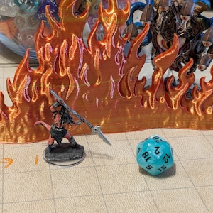 May include: A 3D printed orange and red flame wall miniature accessory for tabletop gaming. The piece features a textured, layered design representing fire. It is displayed alongside a small fantasy warrior figurine, a multi-eyed creature, and a blue 20-sided die.