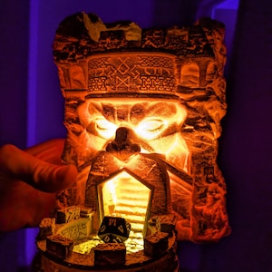 May include: A stone sculpture of a king's head with a glowing orange light inside. The sculpture is a dice tower with a black and blue dice inside. The dice tower is on a wooden base.