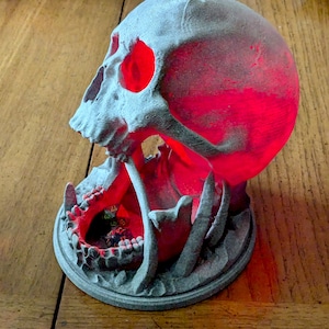 May include: A 3D printed skull dice tower with a red glow emanating from the eye sockets. The tower is designed to hold dice in the mouth of the skull.
