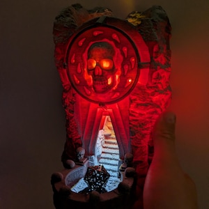 Puede incluir: Una escultura decorativa iluminada con tem&aacute;tica de calaveras. La escultura presenta una calavera dentro de un marco circular, que emite un brillo rojo. El dise&ntilde;o incluye una textura similar a la piedra y una peque&ntilde;a escalera que conduce a una cueva. Una mano sostiene la escultura.