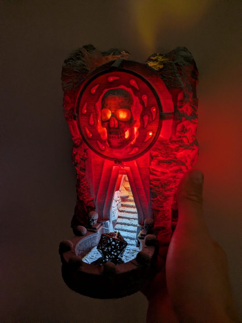 Puede incluir: Una escultura decorativa iluminada con tem&aacute;tica de calaveras. La escultura presenta una calavera dentro de un marco circular, que emite un brillo rojo. El dise&ntilde;o incluye una textura similar a la piedra y una peque&ntilde;a escalera que conduce a una cueva. Una mano sostiene la escultura.