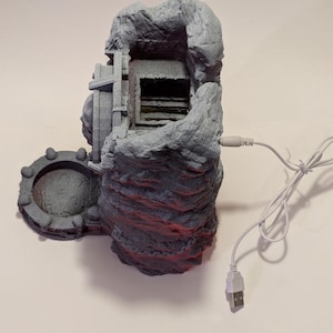 Puede incluir: Una estructura gris en forma de roca con un cable USB adjunto. La estructura tiene una abertura en forma de cueva y una caracter&iacute;stica circular en forma de puerta en el lateral. Se ven detalles en rojo en la parte inferior.