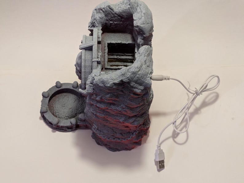 Puede incluir: Una estructura gris en forma de roca con un cable USB adjunto. La estructura tiene una abertura en forma de cueva y una caracter&iacute;stica circular en forma de puerta en el lateral. Se ven detalles en rojo en la parte inferior.