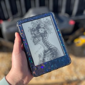 May include: A black e-reader with a decorative, iridescent blue case. The screen shows a black and white illustration of a human skull and spine with floral accents. The case is embellished with small, dark rhinestones.