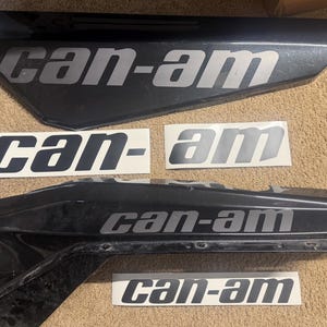 May include: Two black, angular vehicle body panels featuring the silver text can-am, accompanied by four separate adhesive decals, each displaying the silver text can-am on a white rectangular background, arranged in a flat, organized layout.