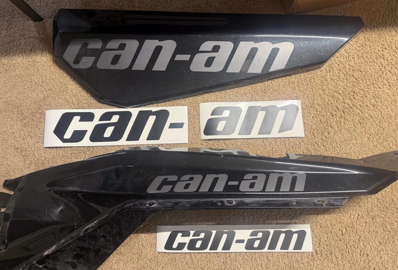 May include: Two black, angular vehicle body panels featuring the silver text can-am, accompanied by four separate adhesive decals, each displaying the silver text can-am on a white rectangular background, arranged in a flat, organized layout.