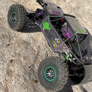 Op de afbeelding: Een zwarte en paarse op afstand bestuurbare rock crawler met grote, noppenbanden en groene accenten. Het voertuig heeft een rolkooi en een doorzichtige carrosserie met afbeeldingen. De banden hebben groene velgen. De crawler staat op een rotsachtig oppervlak.