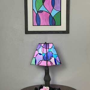 Peut inclure: Une lampe en verre color&eacute; avec un motif g&eacute;om&eacute;trique compos&eacute; de cercles roses, bleus et verts. La lampe est pos&eacute;e sur une table en bois avec une fleur rose et des fleurs noires.
