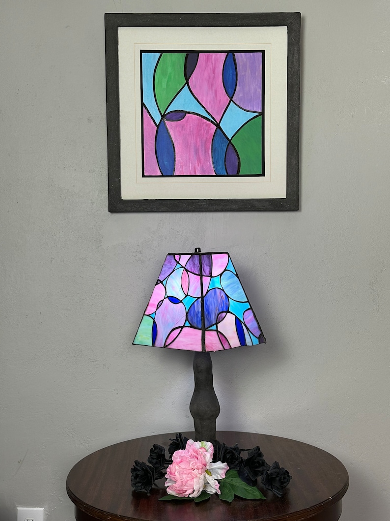 Peut inclure: Une lampe en verre color&eacute; avec un motif g&eacute;om&eacute;trique compos&eacute; de cercles roses, bleus et verts. La lampe est pos&eacute;e sur une table en bois avec une fleur rose et des fleurs noires.