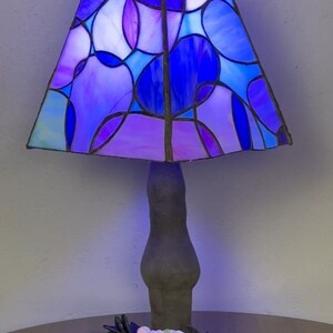 Peut inclure: Une lampe en verre color&eacute; avec une base gris fonc&eacute; et un motif g&eacute;om&eacute;trique bleu, violet et blanc. La lampe est sur une table avec des roses noires et une fleur rose.