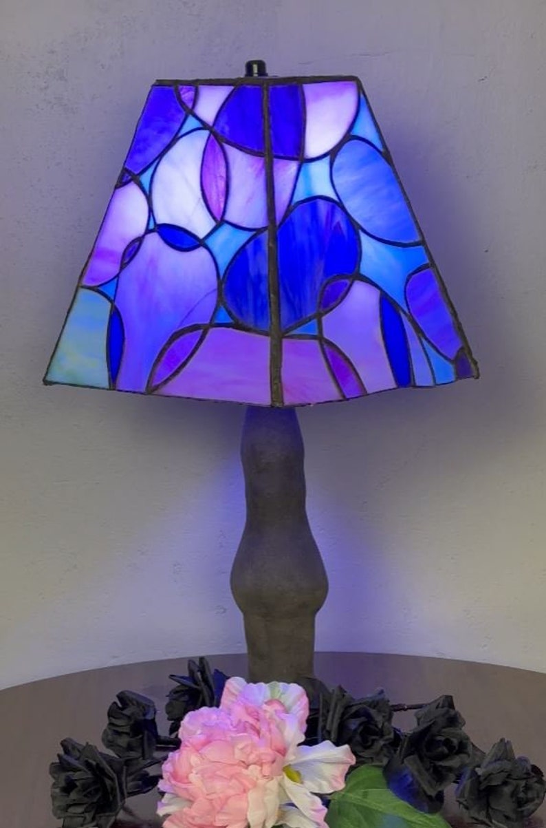 Peut inclure: Une lampe en verre color&eacute; avec une base gris fonc&eacute; et un motif g&eacute;om&eacute;trique bleu, violet et blanc. La lampe est sur une table avec des roses noires et une fleur rose.