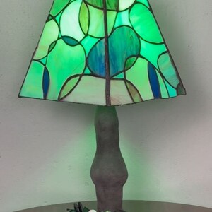 Peut inclure: Une lampe en verre color&eacute; vert et bleu avec un motif g&eacute;om&eacute;trique. La lampe a une base brun fonc&eacute; et est pos&eacute;e sur une table avec des roses noires et une fleur rose.