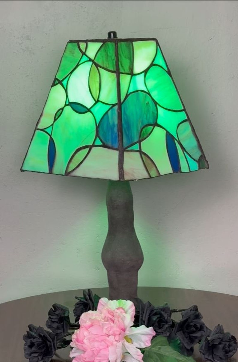Peut inclure: Une lampe en verre color&eacute; vert et bleu avec un motif g&eacute;om&eacute;trique. La lampe a une base brun fonc&eacute; et est pos&eacute;e sur une table avec des roses noires et une fleur rose.