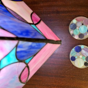 Peut inclure: Deux dessous-de-verre ronds avec un motif en mosa&iuml;que de cercles bleus, violets et verts sur fond blanc. Les dessous-de-verre sont sur une surface en bois brun.