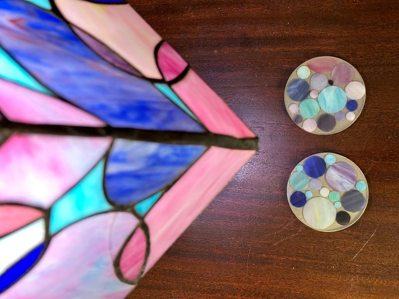 Peut inclure: Deux dessous-de-verre ronds avec un motif en mosa&iuml;que de cercles bleus, violets et verts sur fond blanc. Les dessous-de-verre sont sur une surface en bois brun.
