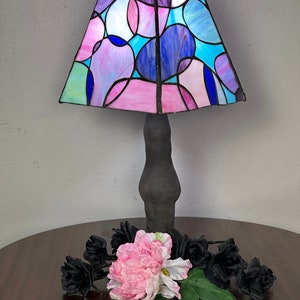 Peut inclure: Une lampe en verre color&eacute; avec une base noire et un motif g&eacute;om&eacute;trique rose, bleu et violet. La lampe est pos&eacute;e sur une table en bois avec des roses noires et une fleur rose.