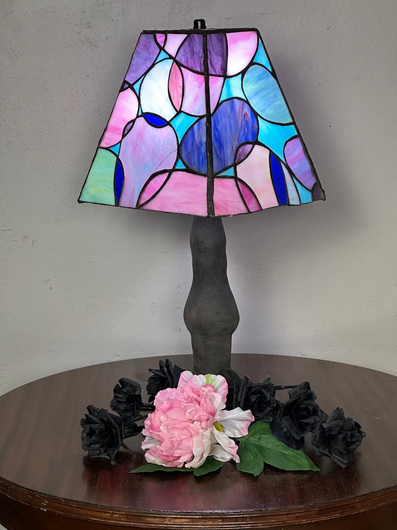 Peut inclure: Une lampe en verre color&eacute; avec une base noire et un motif g&eacute;om&eacute;trique rose, bleu et violet. La lampe est pos&eacute;e sur une table en bois avec des roses noires et une fleur rose.