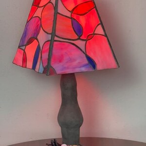 Peut inclure: Une lampe en verre color&eacute; avec un motif g&eacute;om&eacute;trique dans des tons de rose, rouge et bleu. La lampe a une base gris fonc&eacute; et est pos&eacute;e sur une table avec des fleurs noires.