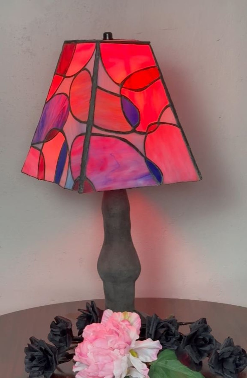Peut inclure: Une lampe en verre color&eacute; avec un motif g&eacute;om&eacute;trique dans des tons de rose, rouge et bleu. La lampe a une base gris fonc&eacute; et est pos&eacute;e sur une table avec des fleurs noires.
