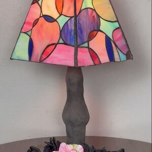 Peut inclure: Une lampe en verre color&eacute; avec une base noire et un motif abstrait color&eacute;. L'abat-jour pr&eacute;sente un motif g&eacute;om&eacute;trique de cercles et d'ovales dans des tons de rose, rouge, bleu, vert et jaune. La lampe est pos&eacute;e sur une table en bois avec des roses noires et une fleur rose.