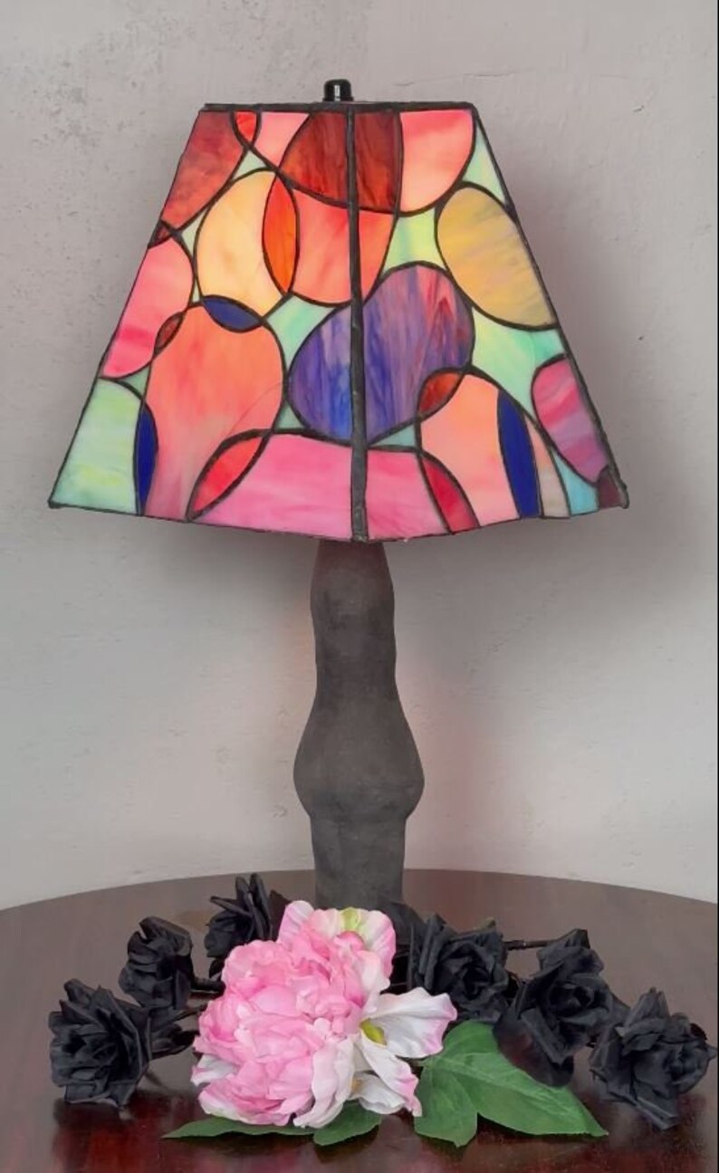 Peut inclure: Une lampe en verre color&eacute; avec une base noire et un motif abstrait color&eacute;. L'abat-jour pr&eacute;sente un motif g&eacute;om&eacute;trique de cercles et d'ovales dans des tons de rose, rouge, bleu, vert et jaune. La lampe est pos&eacute;e sur une table en bois avec des roses noires et une fleur rose.
