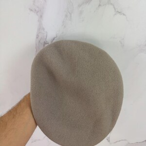 May include: A taupe-colored beret, a classic French-style hat, is held against a marble background. The beret is made of a soft, felt-like material and has a rounded crown. This hat is a versatile accessory for various outfits.