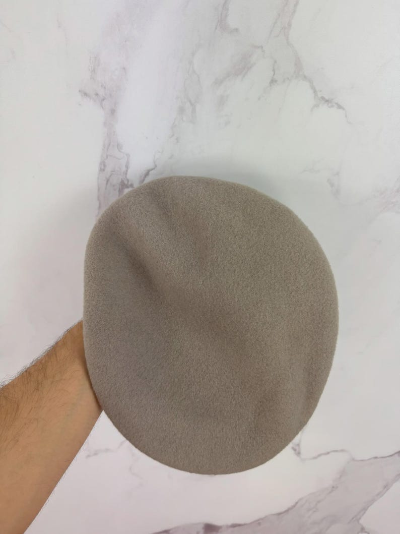 May include: A taupe-colored beret, a classic French-style hat, is held against a marble background. The beret is made of a soft, felt-like material and has a rounded crown. This hat is a versatile accessory for various outfits.