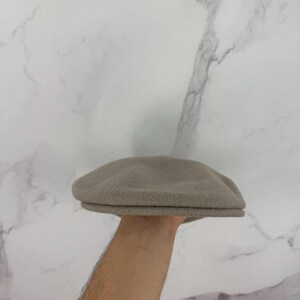 May include: A light brown, wool-like newsboy cap. The cap has a rounded crown and a small, stiff brim. The hat is held up against a white marble background. This classic style hat is suitable for various occasions.