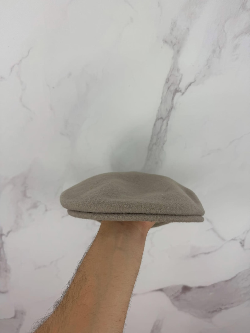 May include: A light brown, wool-like newsboy cap. The cap has a rounded crown and a small, stiff brim. The hat is held up against a white marble background. This classic style hat is suitable for various occasions.