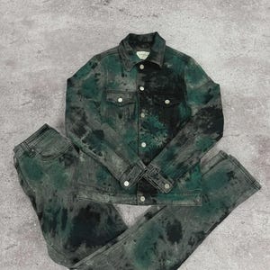 May include: A matching denim jacket and jeans set. The jacket has a button-up front and two chest pockets. The set is dyed in shades of green and black, creating a tie-dye effect. The jeans are straight-legged.