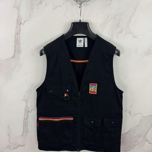 Könnte beinhalten: Schwarze Weste mit Reißverschluss, mehreren Taschen und einem farbenfrohen Bergaufnäher. Die Weste hat ein kleines Adidas-Logo und eine dekorative Verzierung mit orangefarbenen, lila und gelben Akzenten. Die West ist an einem schwarzen Kleiderbügel aufgehängt.