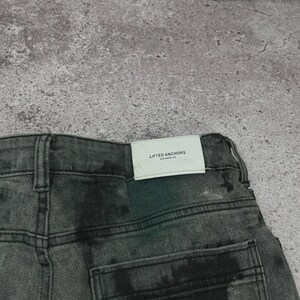 May include: Close-up of the back of a pair of gray denim jeans with a faded, distressed appearance. The jeans have a classic five-pocket design and a white tag that reads "LIFTED ANCHORS LOS ANGELES". The denim has a subtle, mottled pattern.