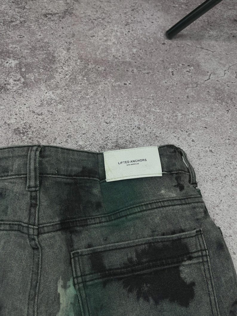 May include: Close-up of the back of a pair of gray denim jeans with a faded, distressed appearance. The jeans have a classic five-pocket design and a white tag that reads "LIFTED ANCHORS LOS ANGELES". The denim has a subtle, mottled pattern.