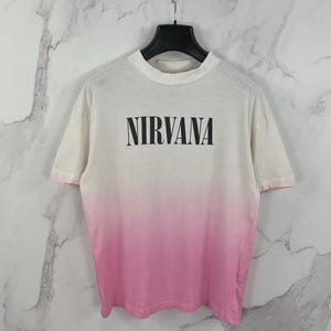 以下が含まれることがあります： 白からピンクへのグラデーションデザインが特徴の半袖クルーネックTシャツ。胸元には黒い大文字で「NIRVANA」という文字がプリントされています。衣類は金属製のチェーンから吊るされた黒いハンガーに掛けられています。
