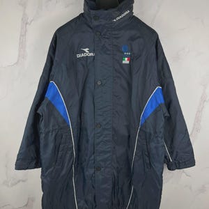 May include: Navy blue Diadora jacket with a high collar and button closure. Features blue and white accents on the sleeves and the Diadora logo on the chest. Includes an Italian flag patch. A vintage, sporty outerwear piece.
