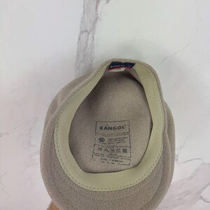 May include: A light brown Kangol fleece hat. The hat has a beige interior band and a tag with the Kangol logo and care instructions. The hat is a classic style, suitable for various occasions.