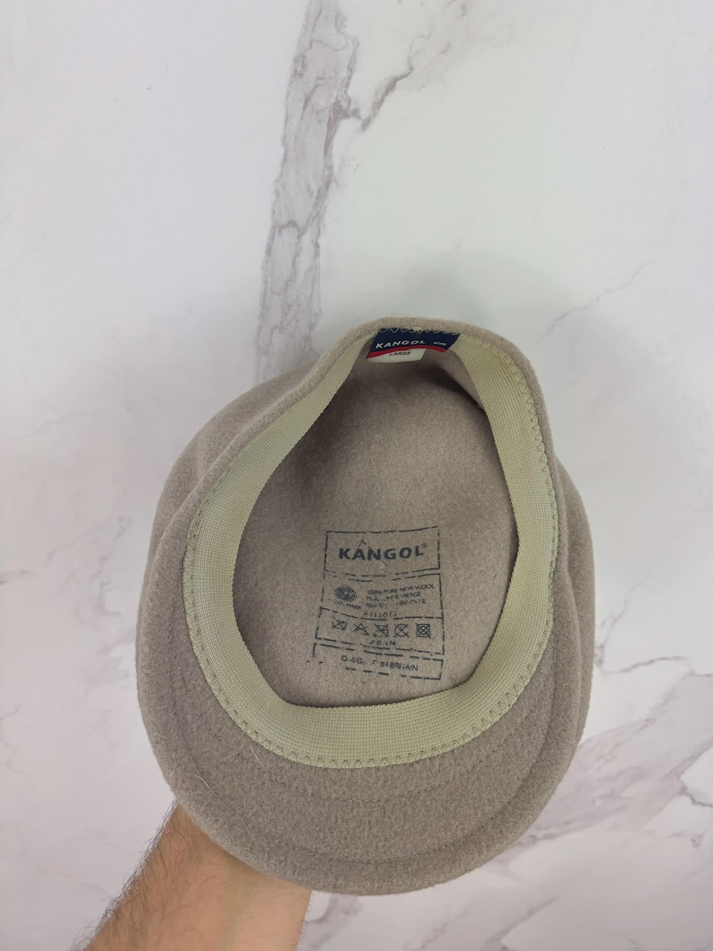 May include: A light brown Kangol fleece hat. The hat has a beige interior band and a tag with the Kangol logo and care instructions. The hat is a classic style, suitable for various occasions.