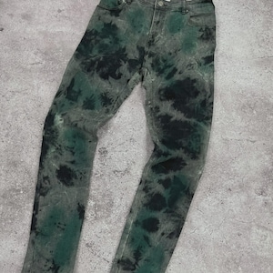 May include: A pair of denim jeans with a unique tie-dye pattern. The jeans feature a mix of green and black dyes on a gray background. The jeans have a classic five-pocket design and a button and zipper closure.