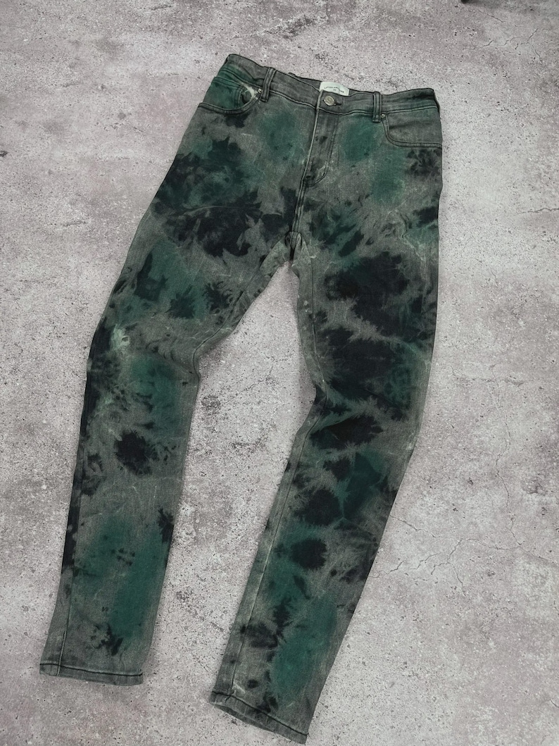 May include: A pair of denim jeans with a unique tie-dye pattern. The jeans feature a mix of green and black dyes on a gray background. The jeans have a classic five-pocket design and a button and zipper closure.