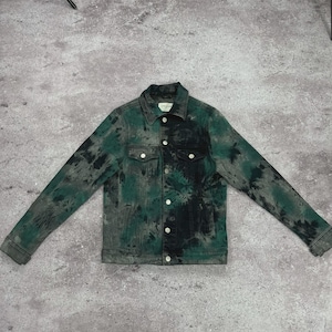 May include: A denim jacket with a tie-dye pattern in shades of green and black. The jacket has a classic design with a button-up front, chest pockets, and long sleeves. The jacket is laid flat on a gray surface.