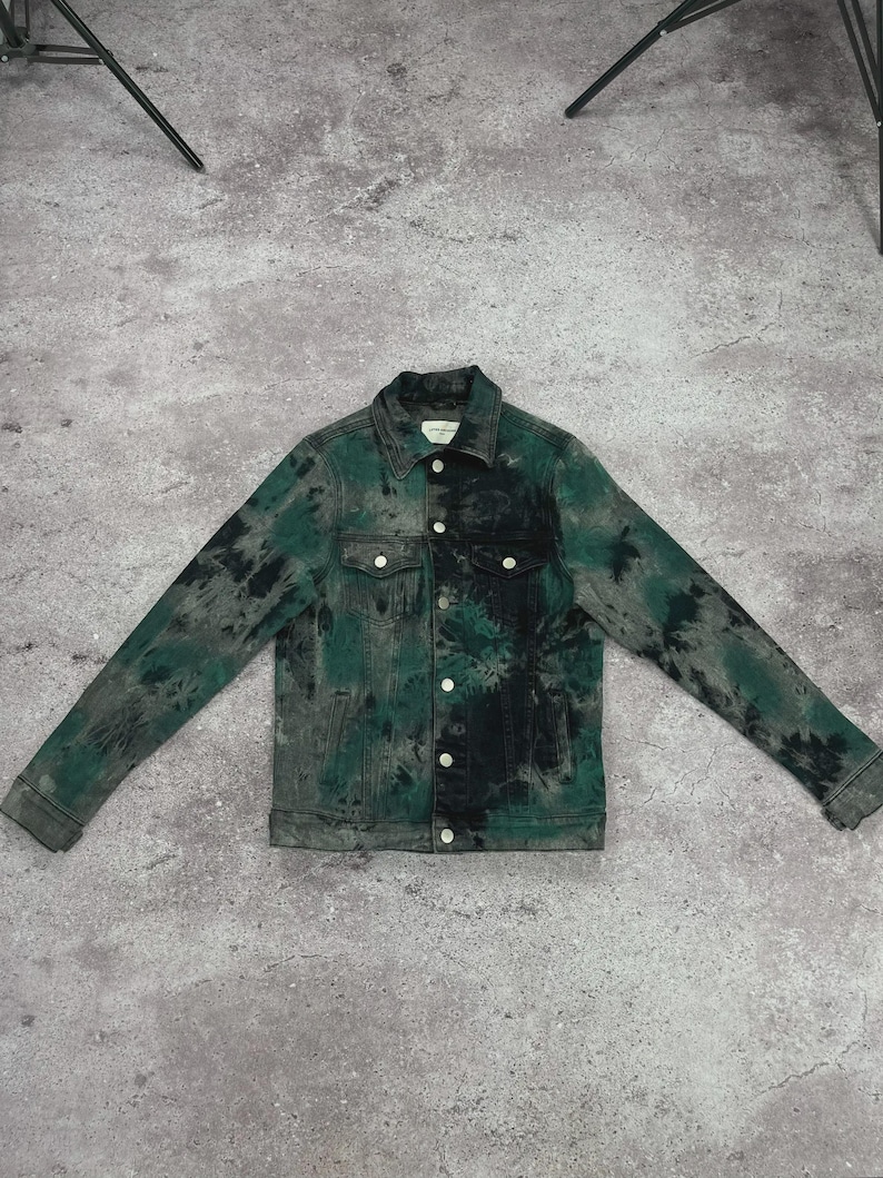 May include: A denim jacket with a tie-dye pattern in shades of green and black. The jacket has a classic design with a button-up front, chest pockets, and long sleeves. The jacket is laid flat on a gray surface.