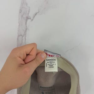 May include: A light brown Kangol wool cap with a beige interior. The cap has a tag that reads "Kangol" and "Large". The cap is made of wool and is a classic style.
